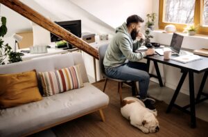 A man works at a desk in a cozy room, focused on his laptop, while a sleeping dog rests beside him on the floor.