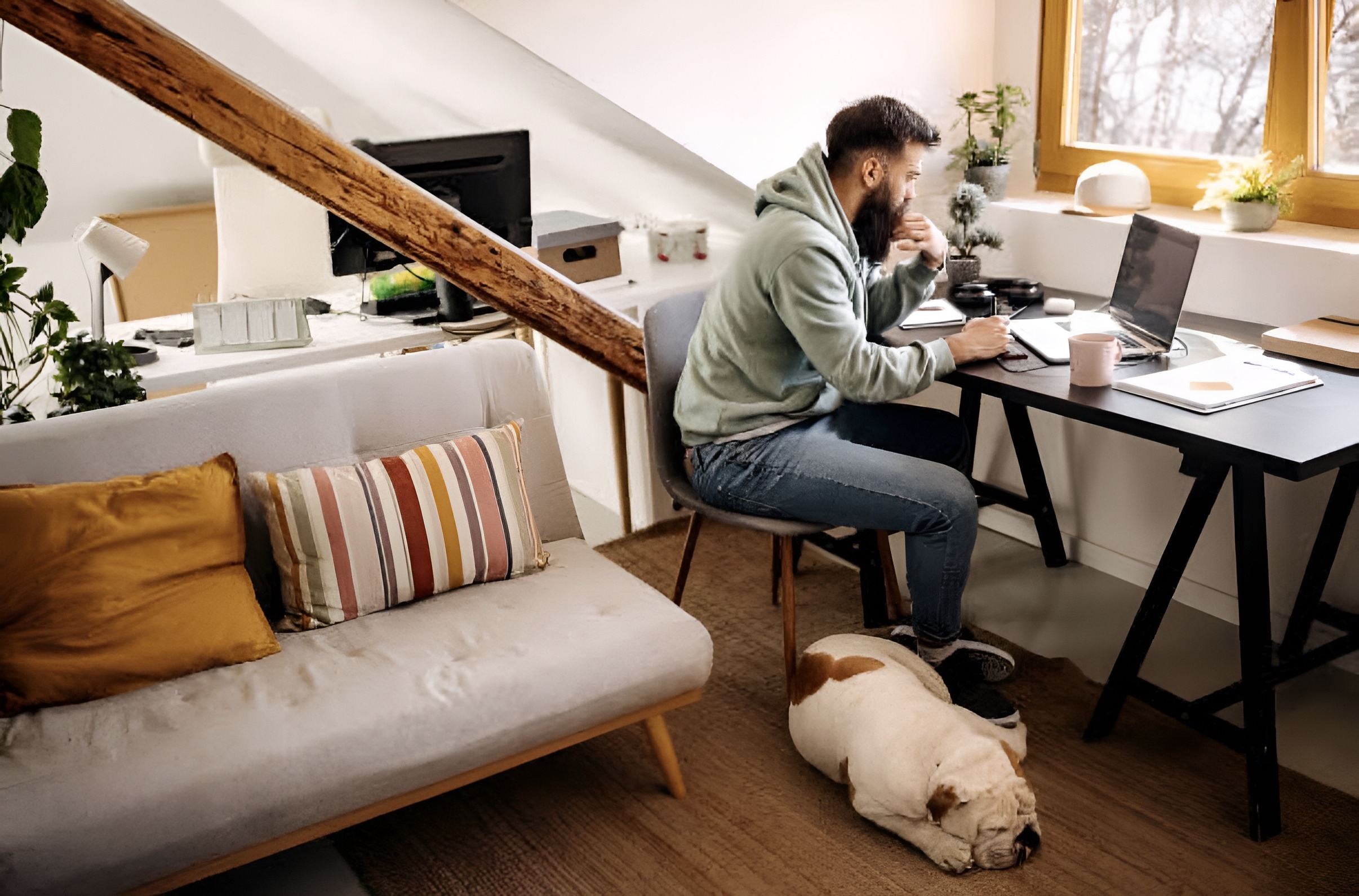 A man works at a desk in a cozy room, focused on his laptop, while a sleeping dog rests beside him on the floor.